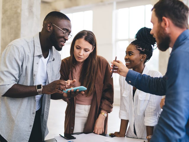 Eine Gruppe von 4 Leuten unterschiedlicher Nationen spricht miteinander. Zwei Männer, zwei Frauen. Ein Mann zeigt was an seinem Handy.