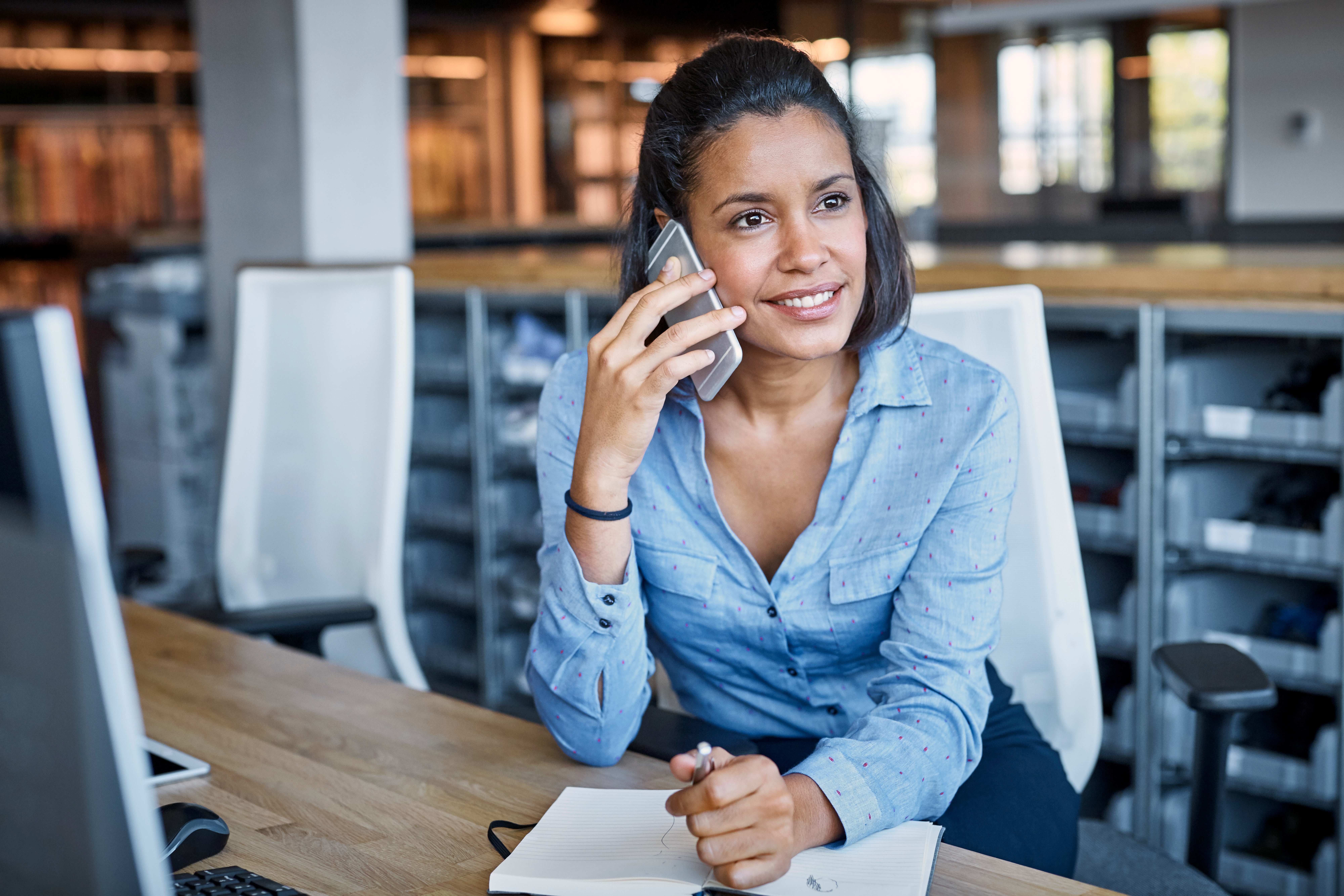 Eine junge Frau sitzt an einem Schreibtisch und telefoniert mit ihrem Handy.