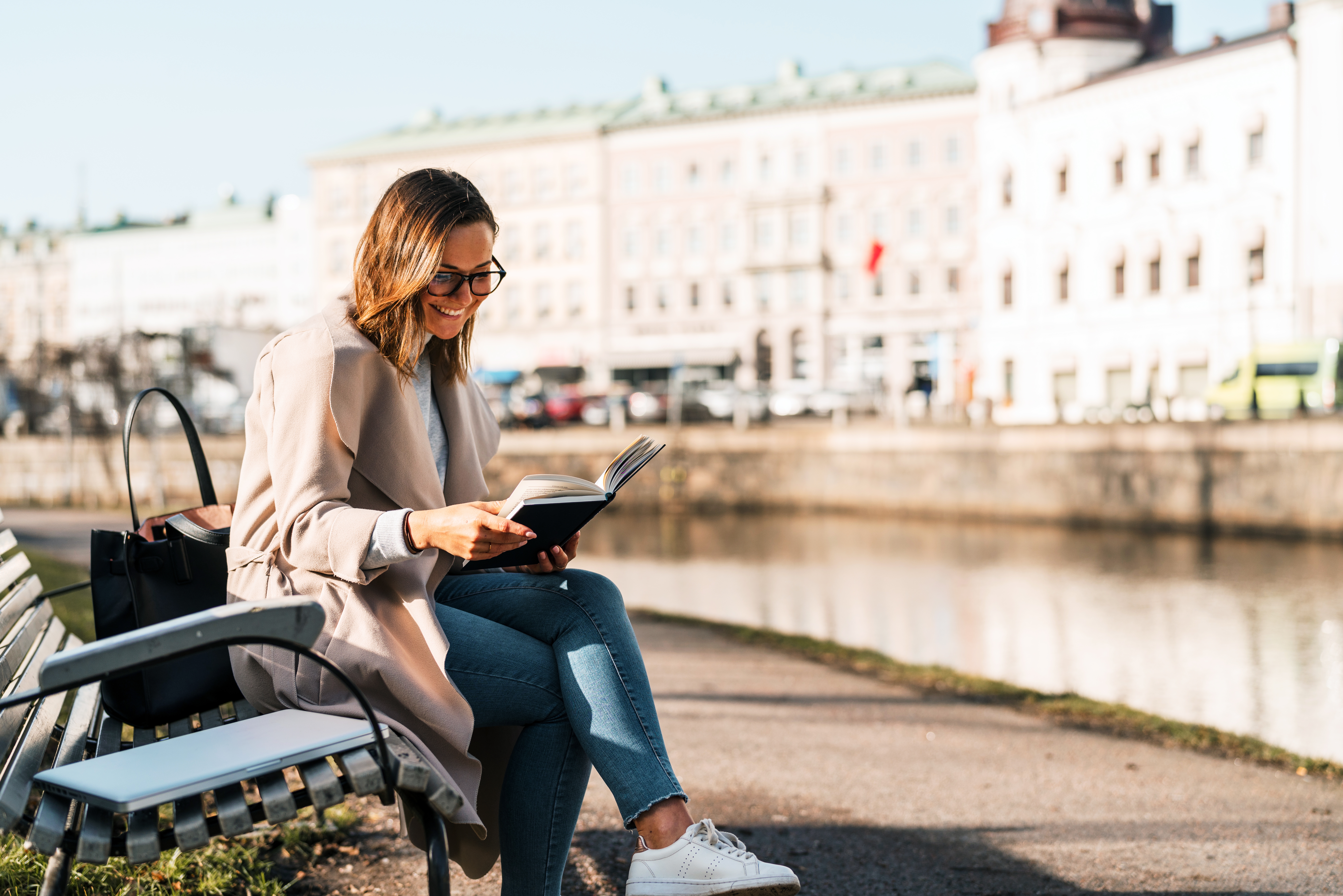 Eine Frau sitzt am Fluss auf einer Bank und liest in einem Buch.