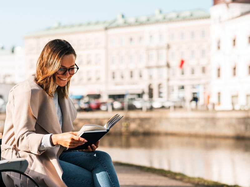 Eine Frau sitzt am Fluss auf einer Bank und liest in einem Buch.
