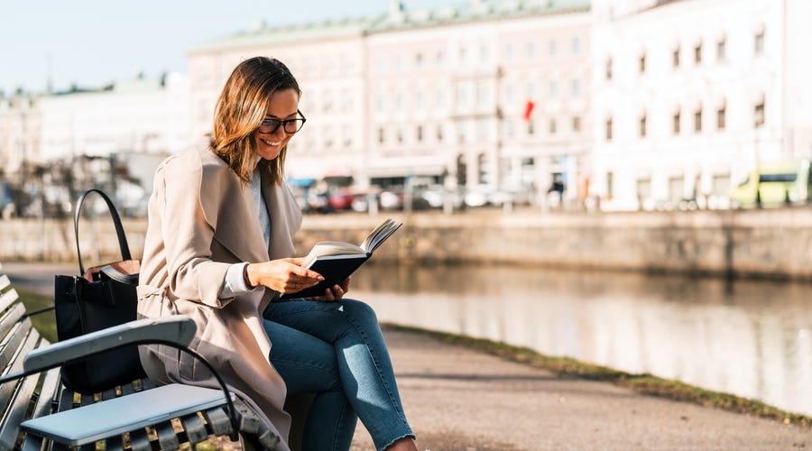 Eine Frau sitzt am Fluss auf einer Bank und liest in einem Buch.