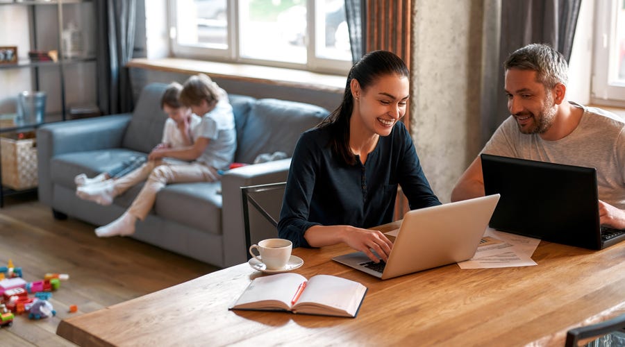 Frau und Mann sitzen am Wohnzimmertisch mit ihrem Laptop. Sie lachen. Ihre kleinen Kinder sitzen hinter ihnen auf dem Sofa.