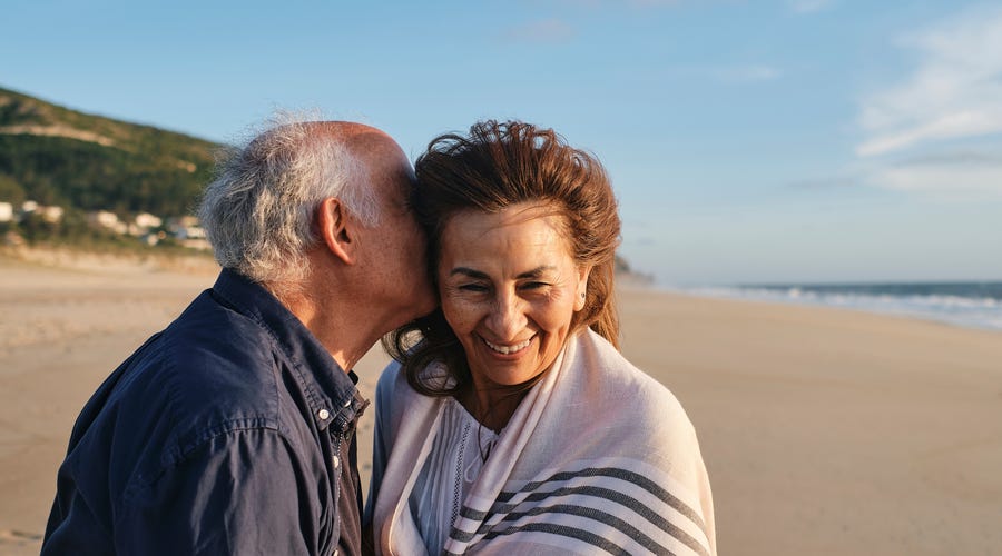 Ein älters Paar, Mann und Frau, am Strand. Er hat küsst sie auf ihre rechte Seite. Sie lächelt direkt in die Kamera.