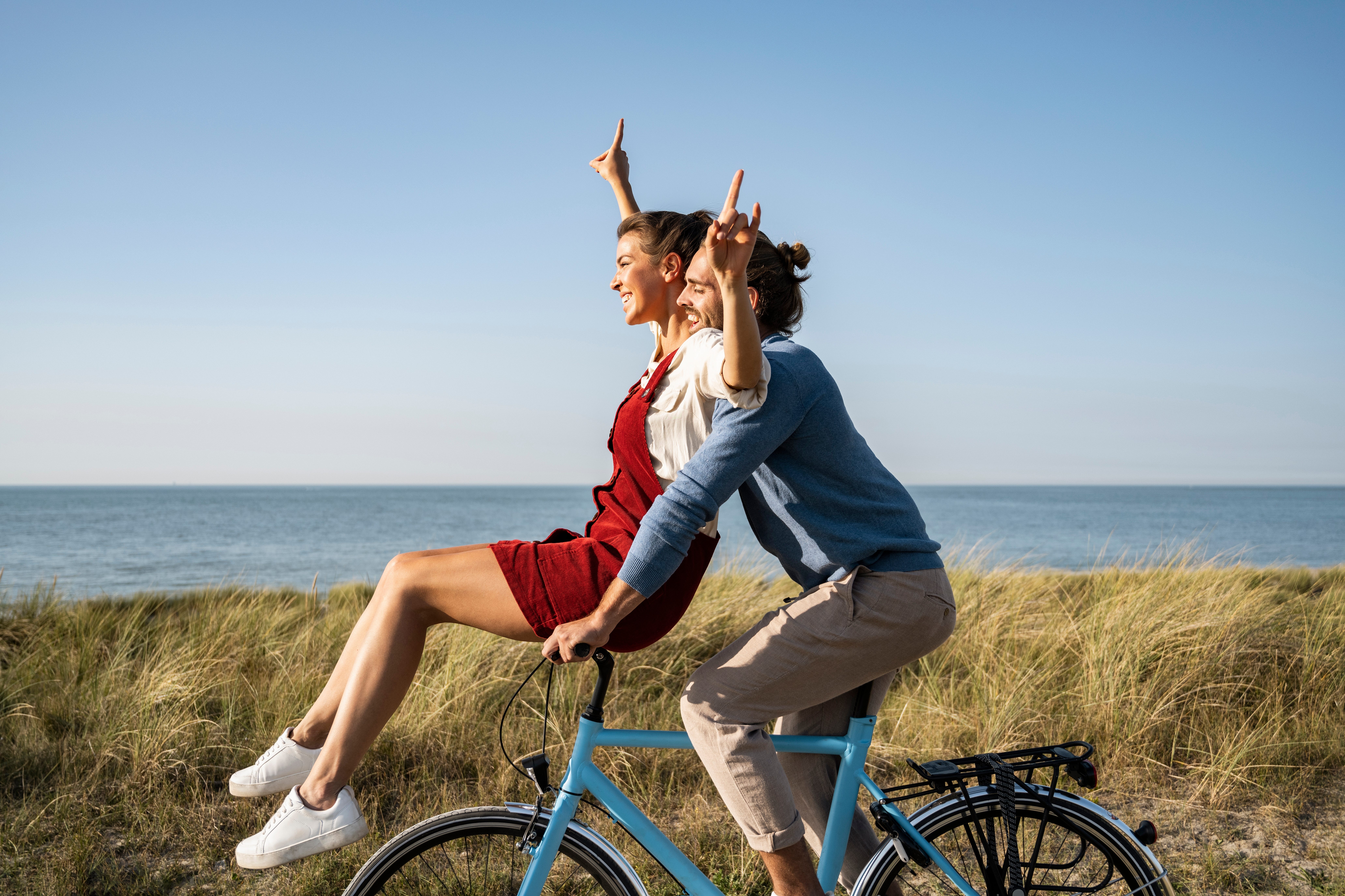 Zwei Personen sitzen auf einem Fahrrad am Meer und lachen.