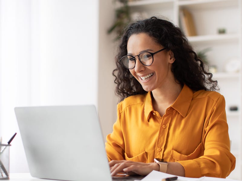 Eine Frau sitzt lächelnd am Laptop. Sie trägt eine gelbe Bluse, eine schwarze Brille, lächelt und hat lockige dunkle Haare.