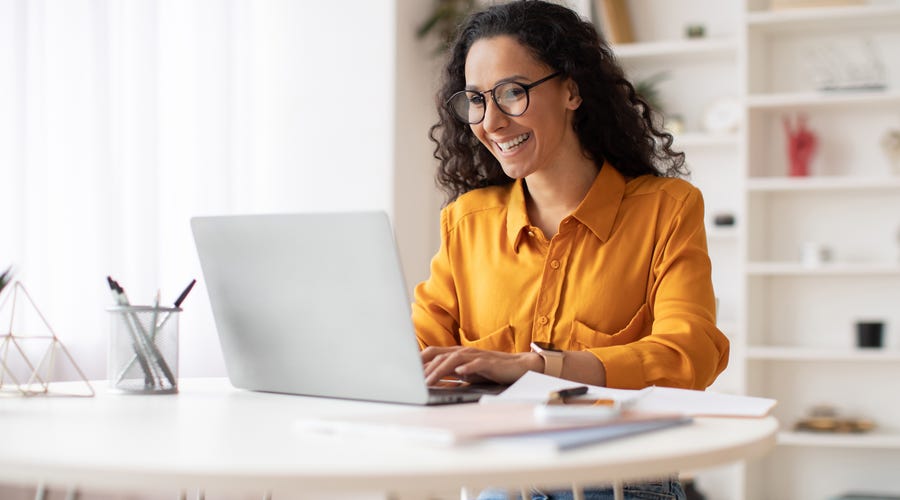 Eine Frau sitzt lächelnd am Laptop. Sie trägt eine gelbe Bluse, eine schwarze Brille, lächelt und hat lockige dunkle Haare.