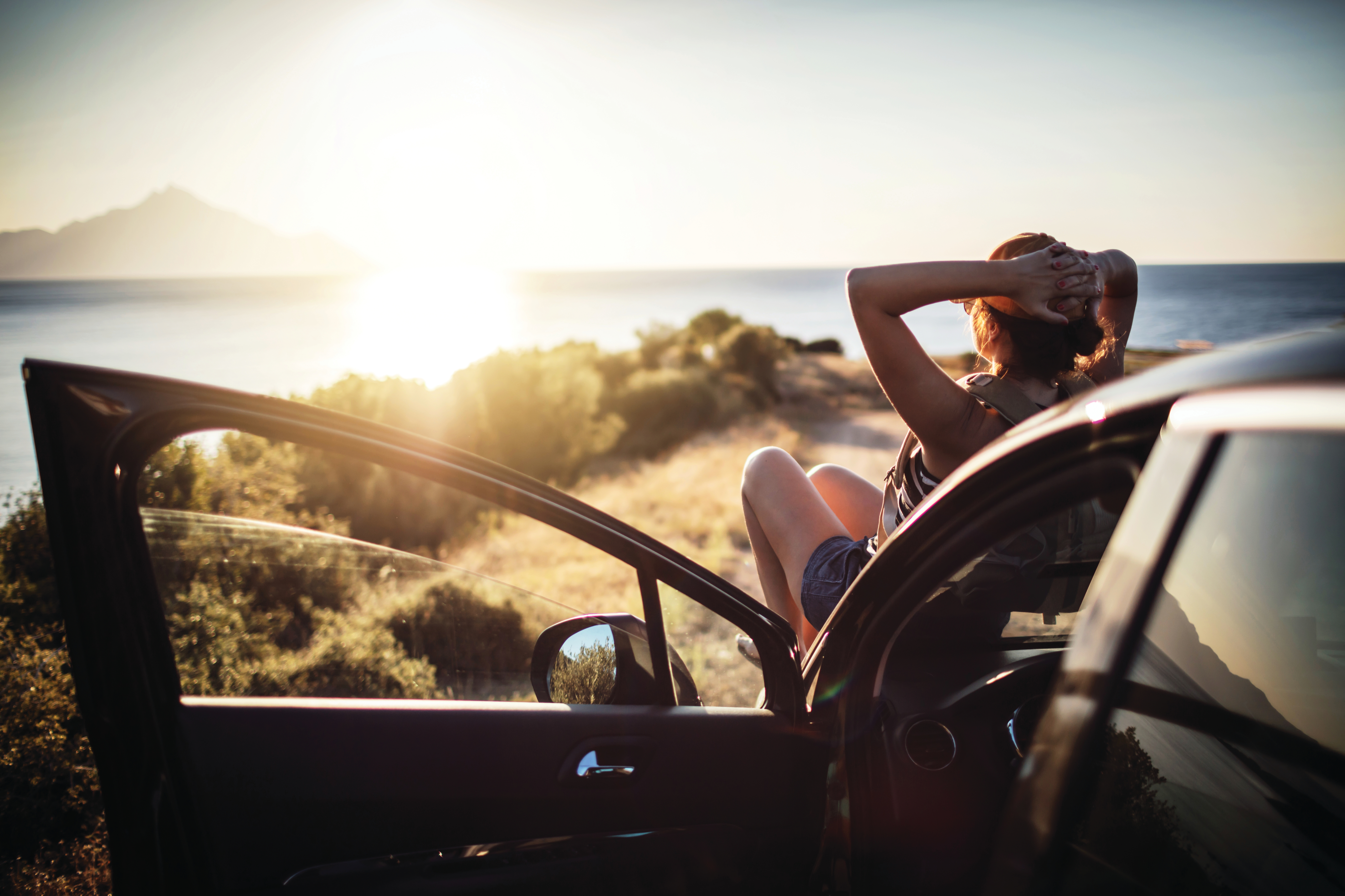 Eine Frau liegt auf einem Auto und schaut der im Meer untergehenden Sonne zu.