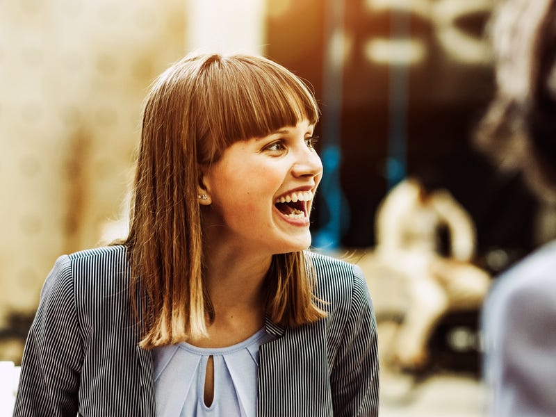 Junge Frau sitzt mit weiteren Personen an einem Tisch und freut sich.