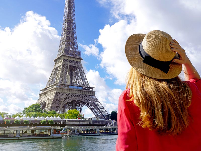 Frau mit Hut steht mit dem Rücken zur Kamera vor dem Eiffelturm.