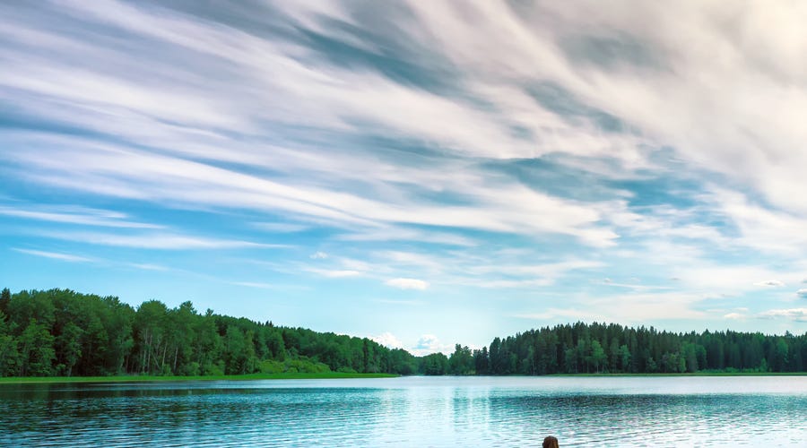 Eine Frau sitzt auf einem Holzsteg am Rande eines Sees und blickt auf das Wasser.