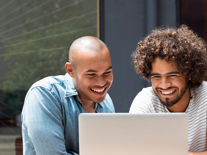2 Männer sitzen vor einem Laptop und lachen.