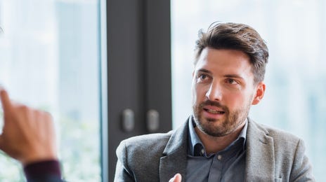 Geschäftsmannn in grauem Anzug erklärt einer anderen Person etwas. Andere Person sieht man nicht.