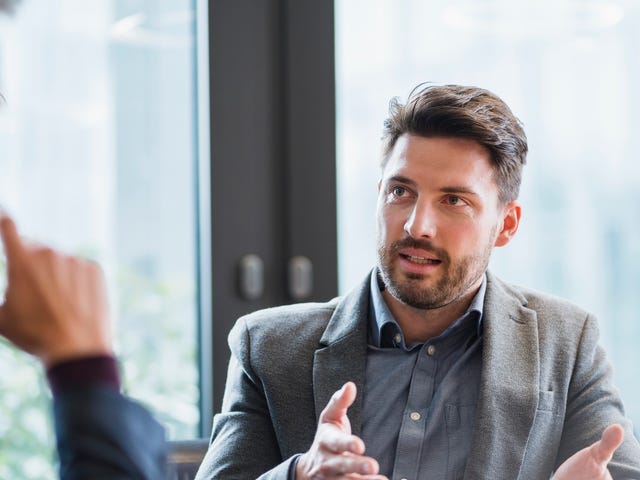 Geschäftsmannn in grauem Anzug erklärt einer anderen Person etwas. Andere Person sieht man nicht.