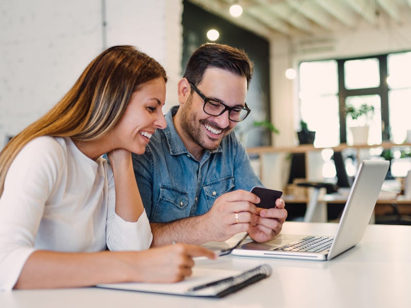 Eine Frau und ein Mann sitzen am Schreibtisch und schauen sich etwas auf dem Smartphone an.