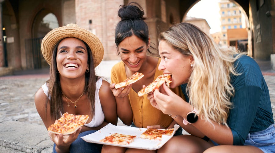 3 junge Frauen sitzen lachend neben einander und essen Pizza.