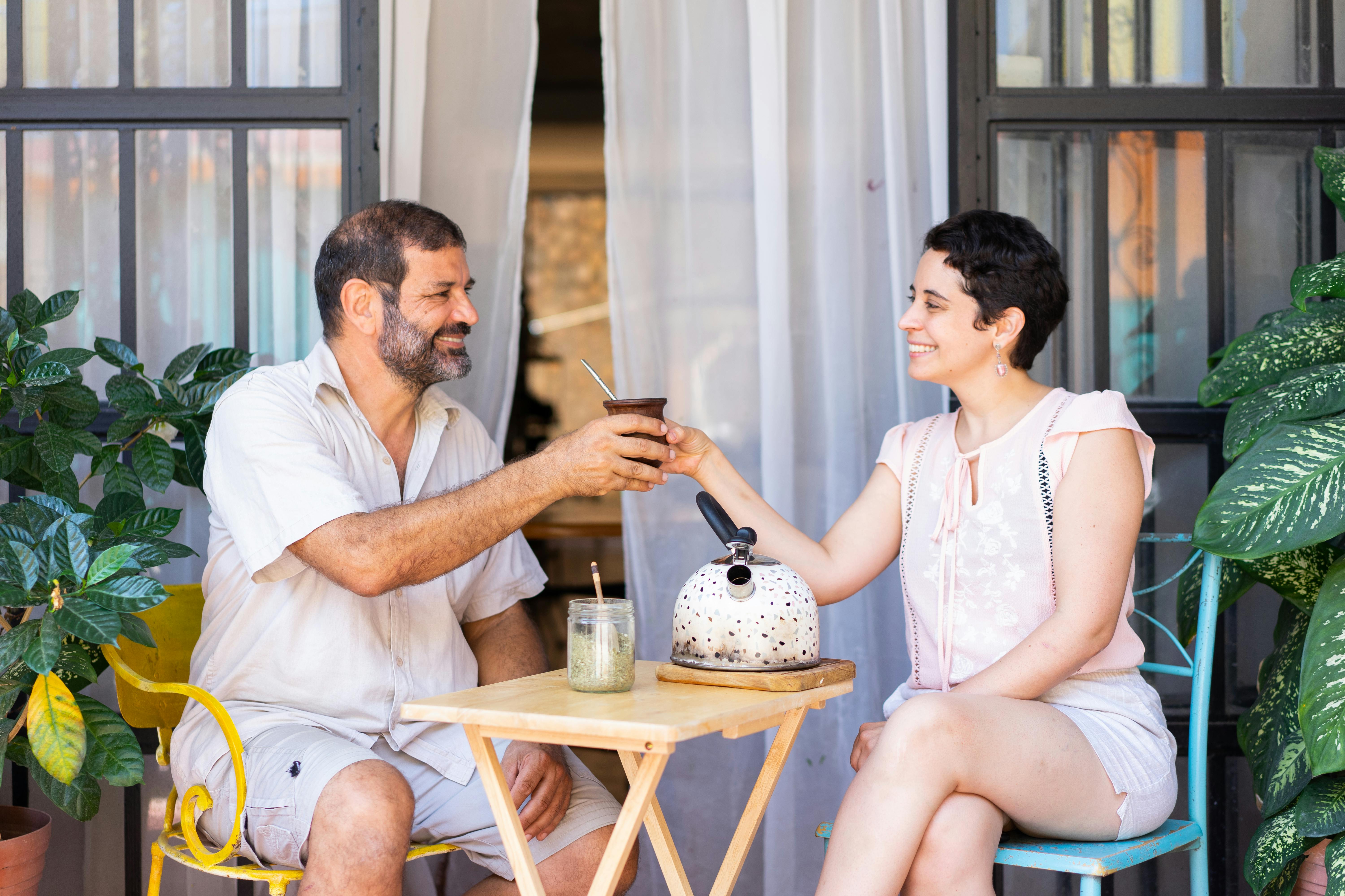 Eine Frau und ein Mann sitzen draußen vor einem Cafe und halten lächelnd ein Becher Mate in der Hand.