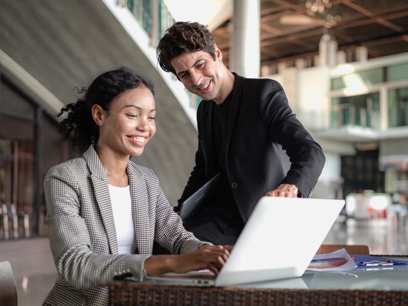 Ein Mann und eine Frau sitzen vor einem Laptop und der Mann zeigt lächelnd auf den Bildschirm.