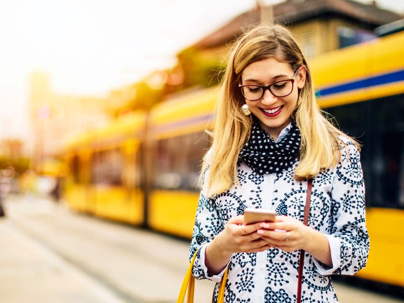 Eine Frau steht an der Haltestelle und tippt eine Nachricht auf ihrem Smartphone.