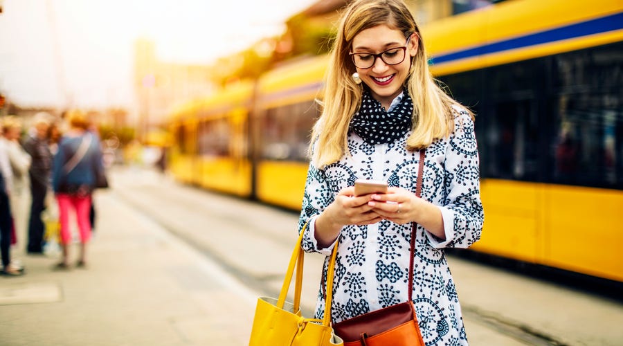 Eine Frau steht an der Haltestelle und tippt eine Nachricht auf ihrem Smartphone.