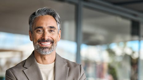 Man in beigem Anzug mit Armen verschränkten Armen schaut in die Kamera und lächelt.