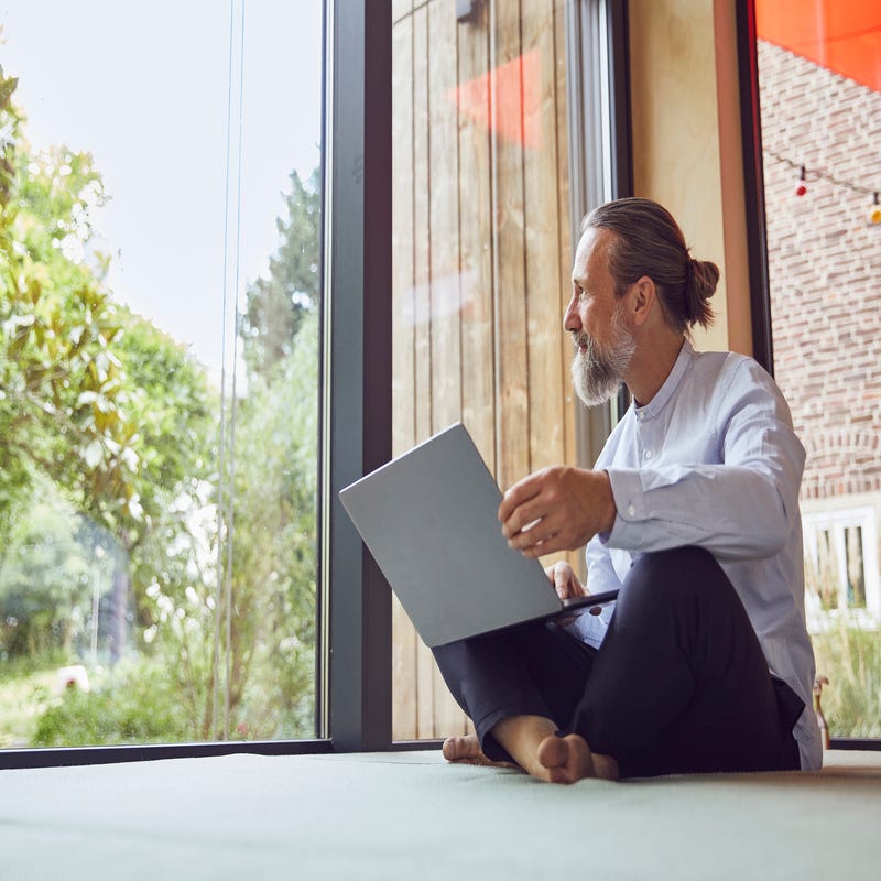 Ein Mann sitzt mit dem Laptop auf dem Schoß vor einem großen Fenster.
