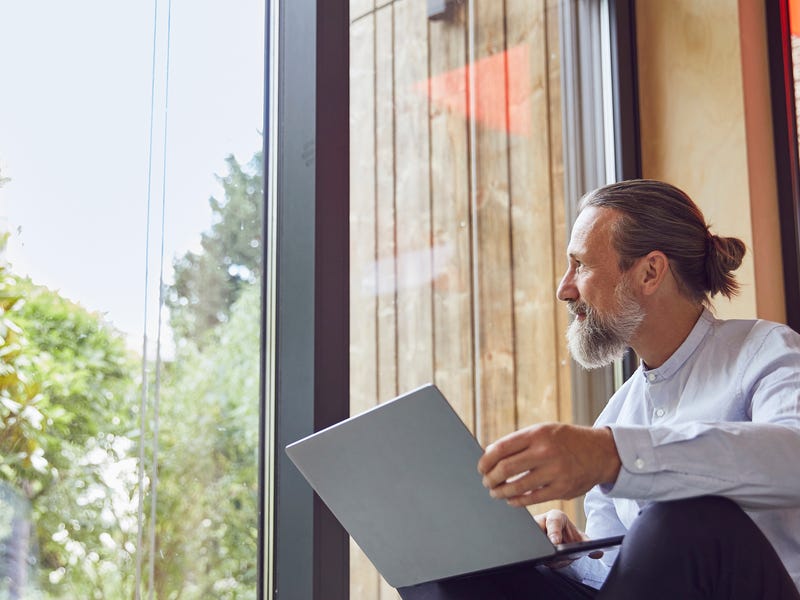 Ein Mann sitzt mit dem Laptop auf dem Schoß vor einem großen Fenster.