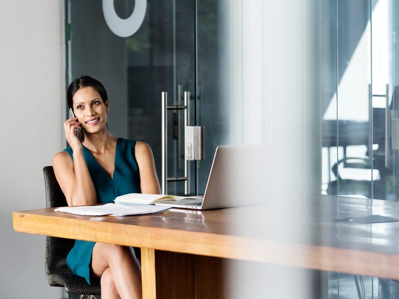 Eine Frau sitzt am Tisch mit Laptop und Arbeitsbüchern und telefoniert.