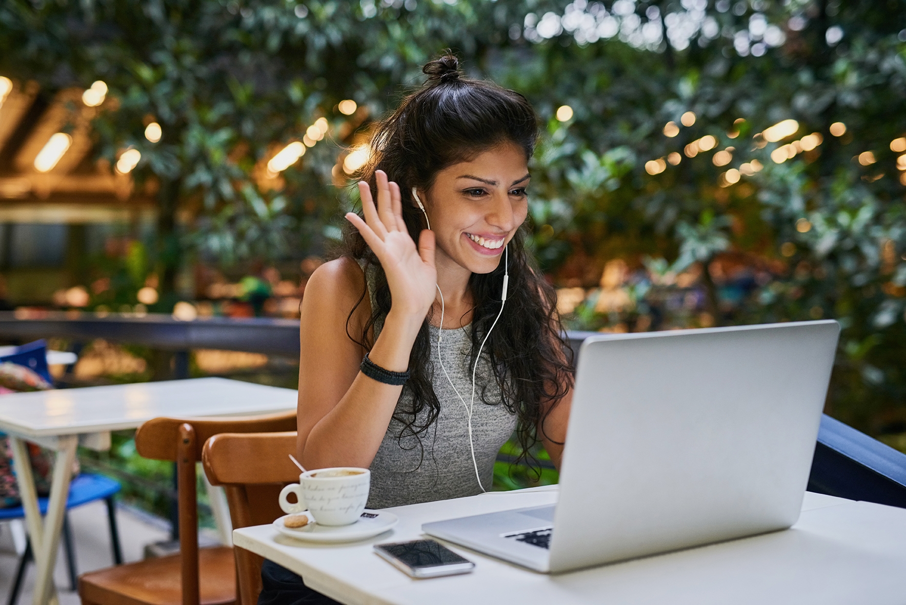 Eine Frau mit ihrem Laptop in einem Café winkt den anderen Teilnehmern ihres Online-Sprachkurses zu.