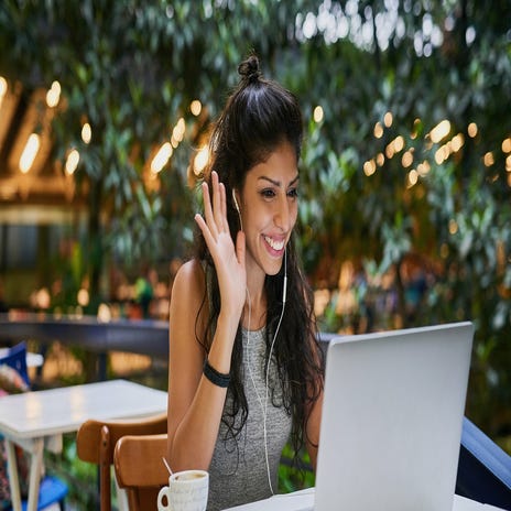 Eine Frau mit ihrem Laptop in einem Café winkt den anderen Teilnehmern ihres Online-Sprachkurses zu.