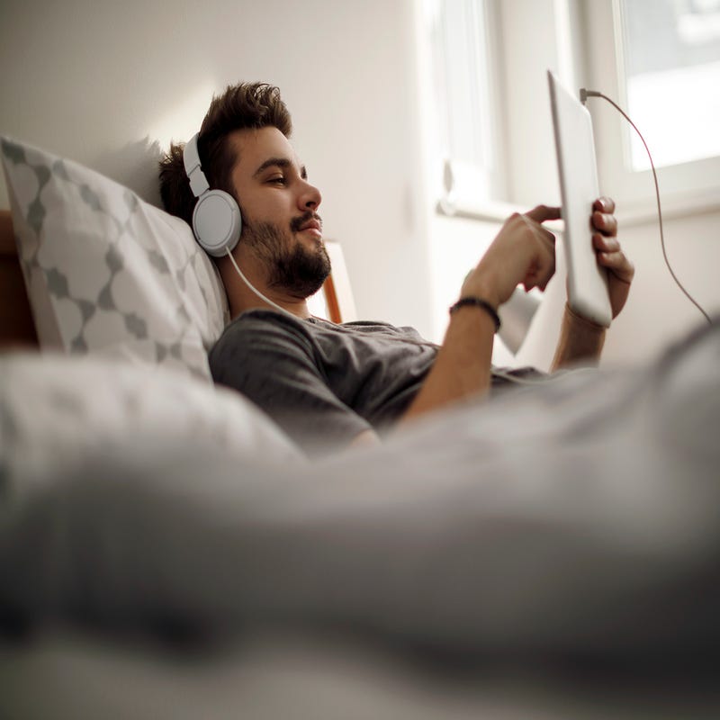 Ein Mann liegt auf dem Bett, mit Tablet und Kopfhörern.