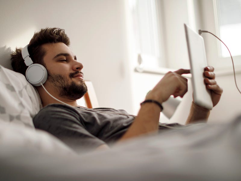 Ein Mann liegt auf dem Bett, mit Tablet und Kopfhörern.