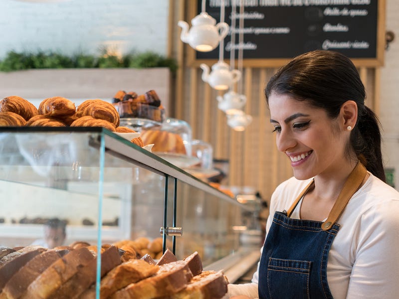 Eine junge Frau steht hinter einer Theke in einem Cafe und zieht ein Tablett mit Kuchen aus der Vitrine.
