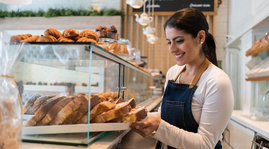 Eine junge Frau steht hinter einer Theke in einem Cafe und zieht ein Tablett mit Kuchen aus der Vitrine.