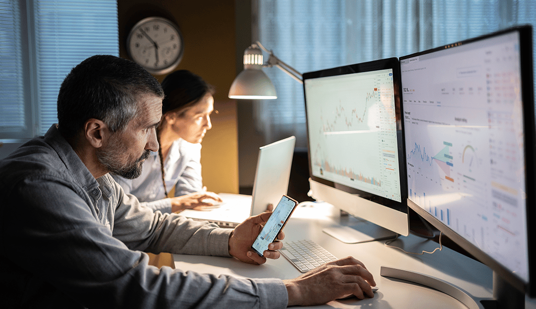 Two People Working at a Shared Desk with Monitors All Around Them