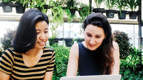Two young women studying together with a laptop in a lush, green setting, representing the concept of learning Thai online in a modern, collaborative environment.