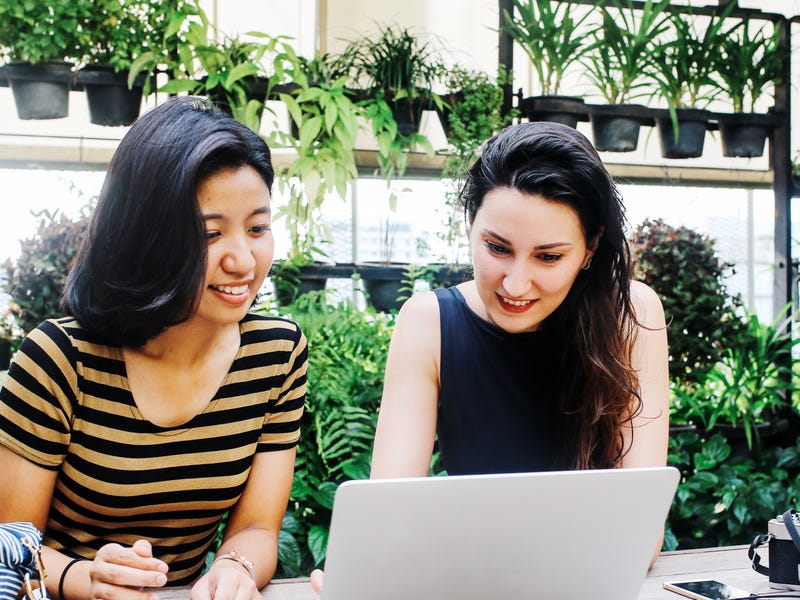Two young women studying together with a laptop in a lush, green setting, representing the concept of learning Thai online in a modern, collaborative environment.
