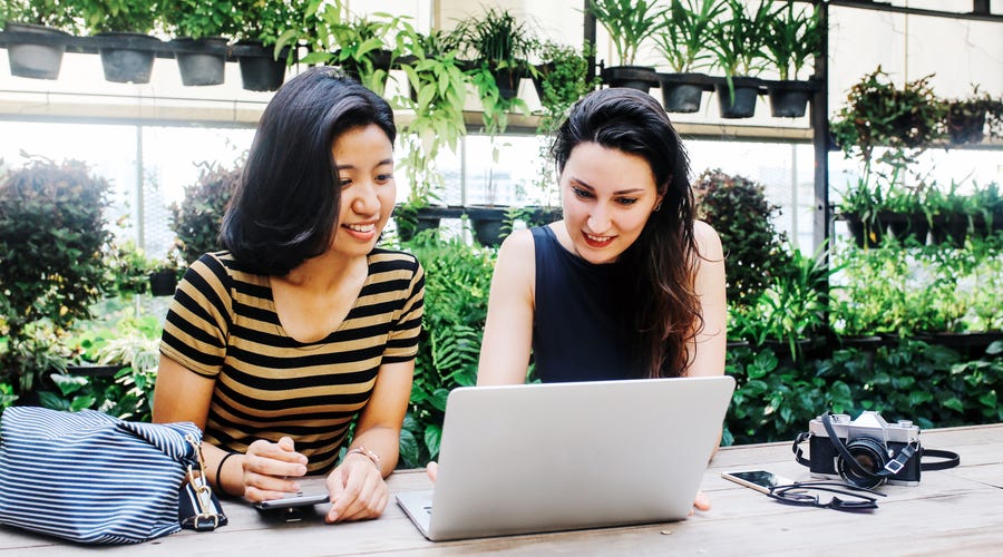 Two young women studying together with a laptop in a lush, green setting, representing the concept of learning Thai online in a modern, collaborative environment.
