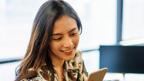 A woman in an office setting, smiling while using her phone, representing a modern approach to global corporate language training.