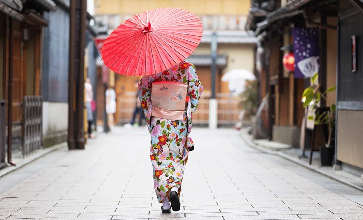 A person wearing a traditional kimono and holding a red parasol walks through a historic Japanese street, symbolizing cultural immersion through Japanese classes.