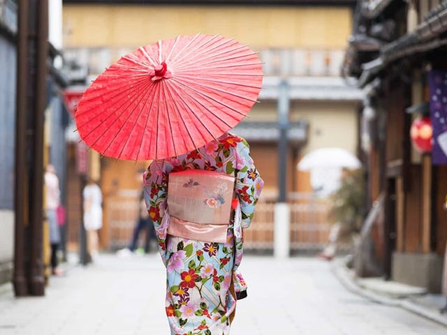 A person wearing a traditional kimono and holding a red parasol walks through a historic Japanese street, symbolizing cultural immersion through Japanese classes.