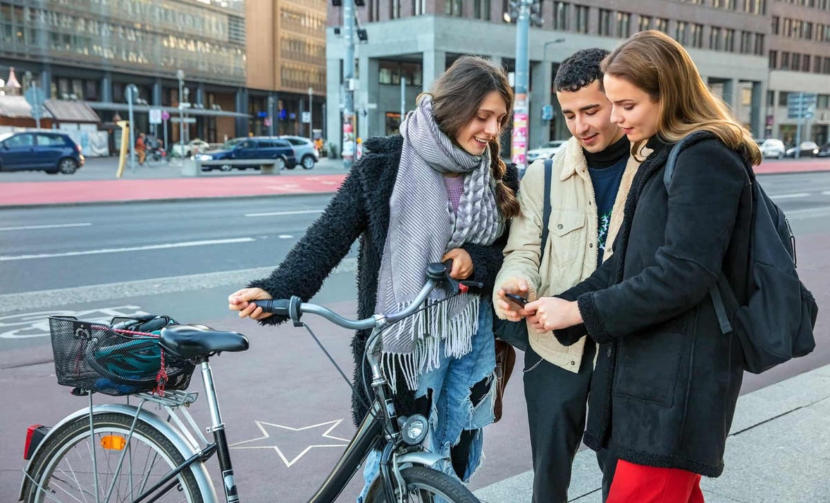 Three young adults smiling and checking a phone together outdoors, representing global connection and community while learning French online.