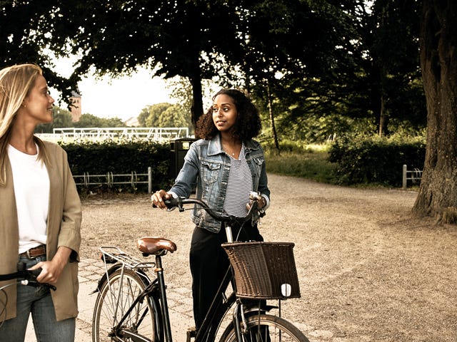 Two women talking casually while walking their bikes after attending Dutch classes near me in a local park setting.