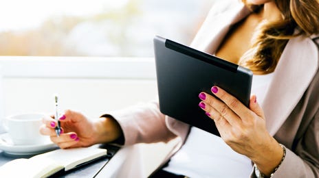 Smiling woman taking a written English test on a tablet while seated at a bright, modern cafe with a notebook and coffee.