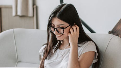 Smiling young woman with glasses using a laptop on a sofa, preparing for a Dutch teaching career online.