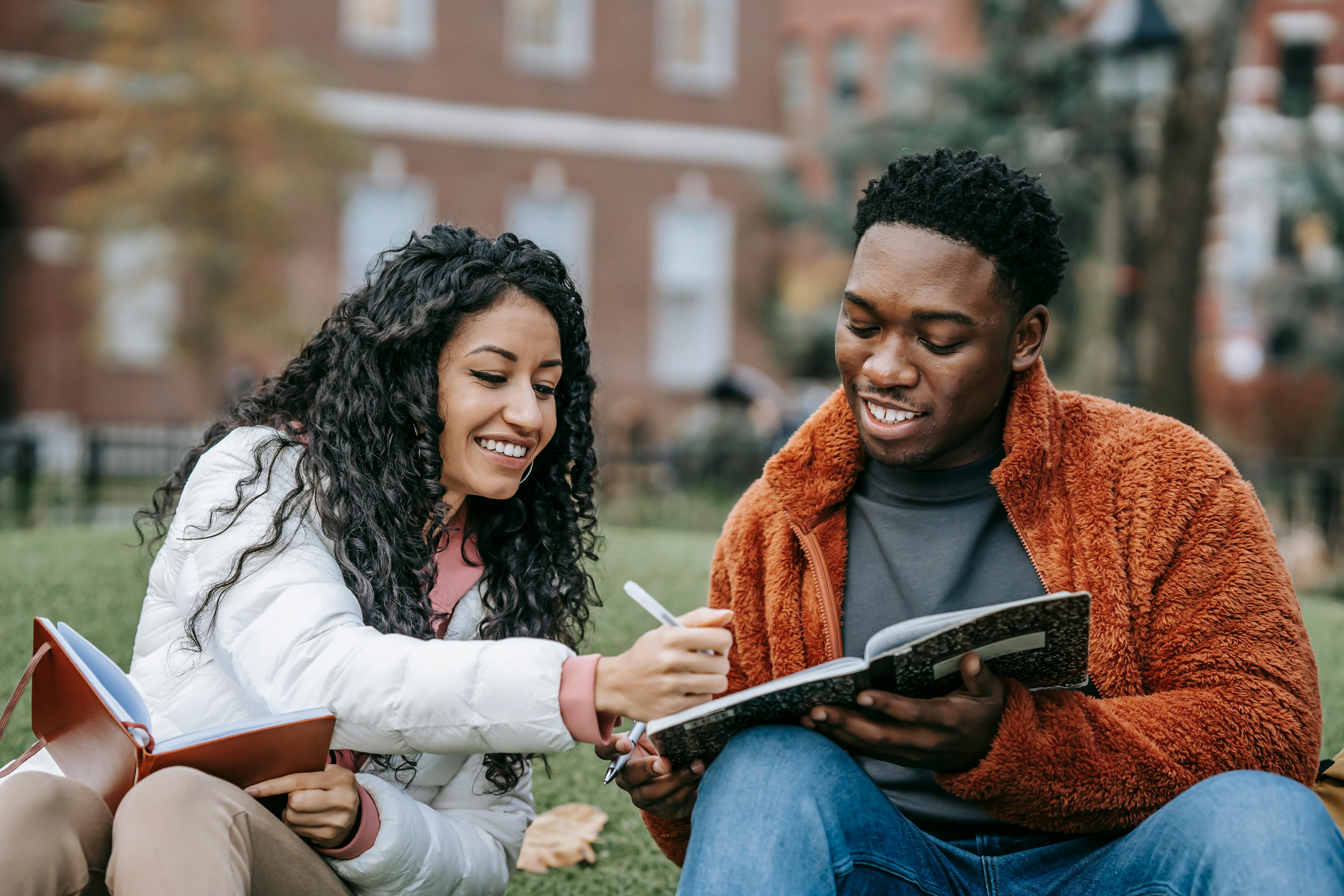 Two friends sitting in a park with their notebooks and practicing the present continuous tense together