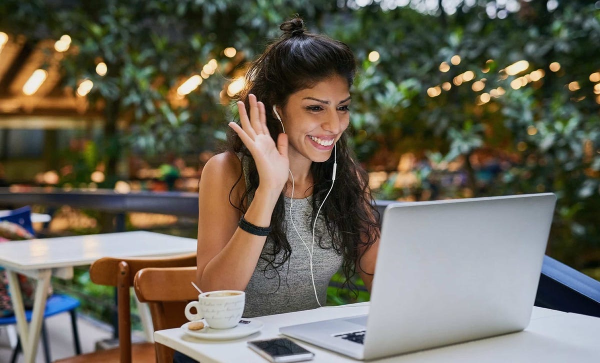 Smiling woman waving during a virtual session at a café, showcasing the accessibility of Berlitz online language schools for global learners.