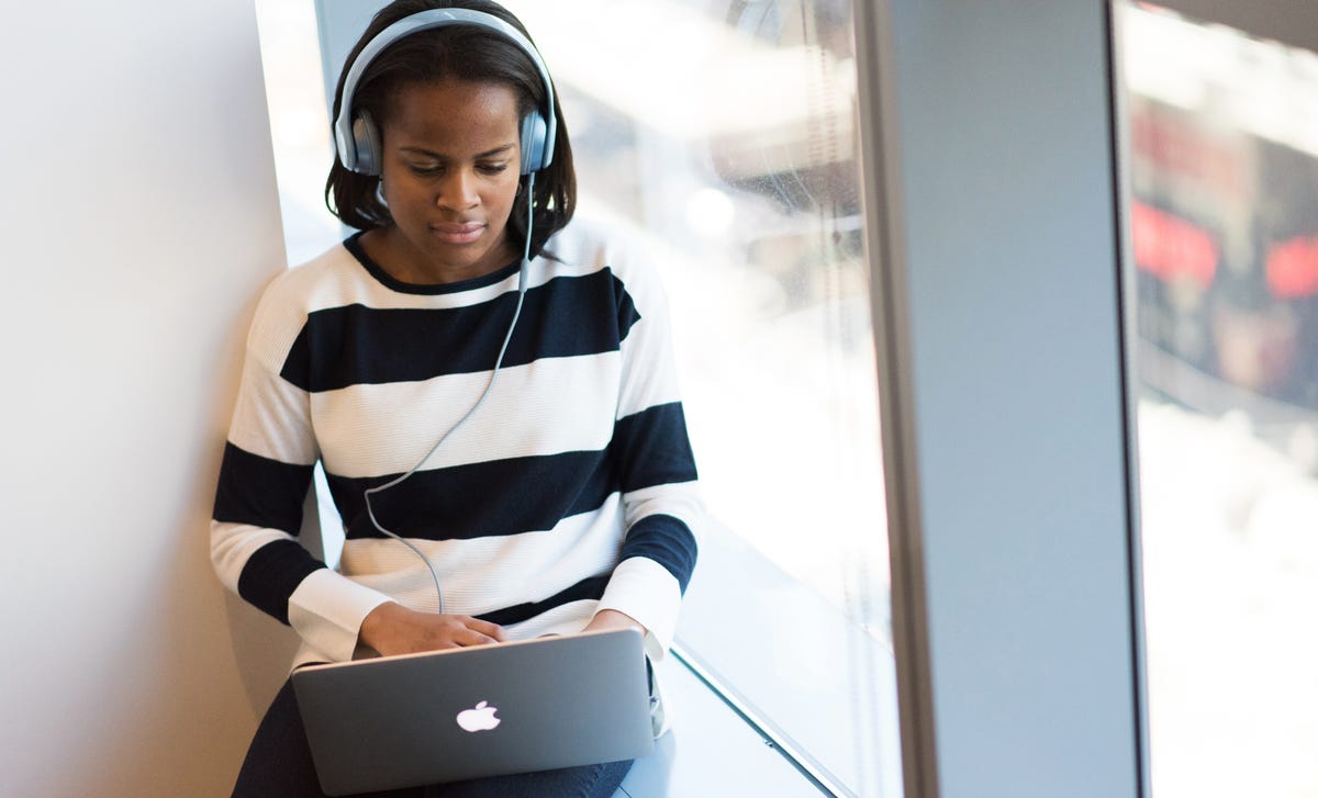Focused young woman wearing headphones and studying on her laptop during an online Korean class by the window.