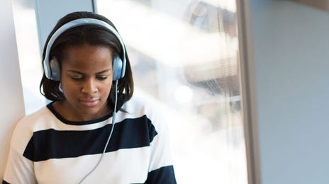 Focused young woman wearing headphones and studying on her laptop during an online Korean class by the window.