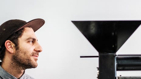A man in a casual cap and button-down shirt working near industrial machinery, representing practical, hands-on ways to learn advanced German in a professional setting.