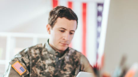 Military man in uniform using a tablet to prepare for the DLPT test in an office with an American flag in the background.